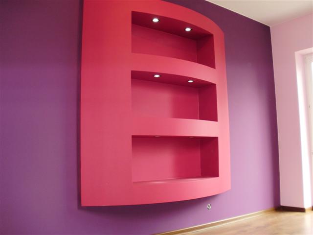 Wnęka ścienna z trzema półkami o zaokrąglonym kształcie, podświetlona od góry małymi lampkami, pomalowana na różowo, na tle fioletowej ściany.